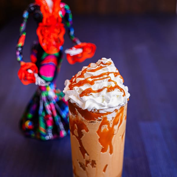 Caramel iced coffee topped with whipped cream, photographed beside a colorful decorative figurine on a dark table.