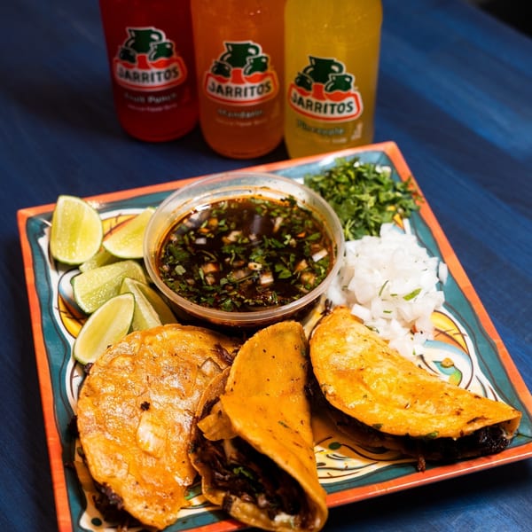Colorful tacos with lime wedges, diced onions, cilantro, and dipping sauce on a decorative plate beside three Jarritos sodas.