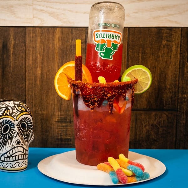 Red chamoy-rimmed drink with fruit garnishes, candy straws, and a mini beer bottle beside a decorative sugar skull.