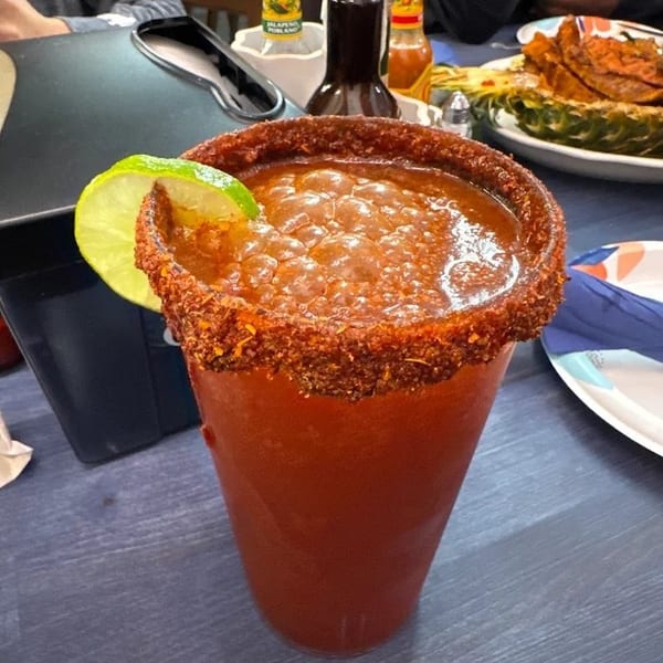 Spicy red cocktail with a chili-seasoned rim and lime wedge on a restaurant table, with condiments and food in the background.