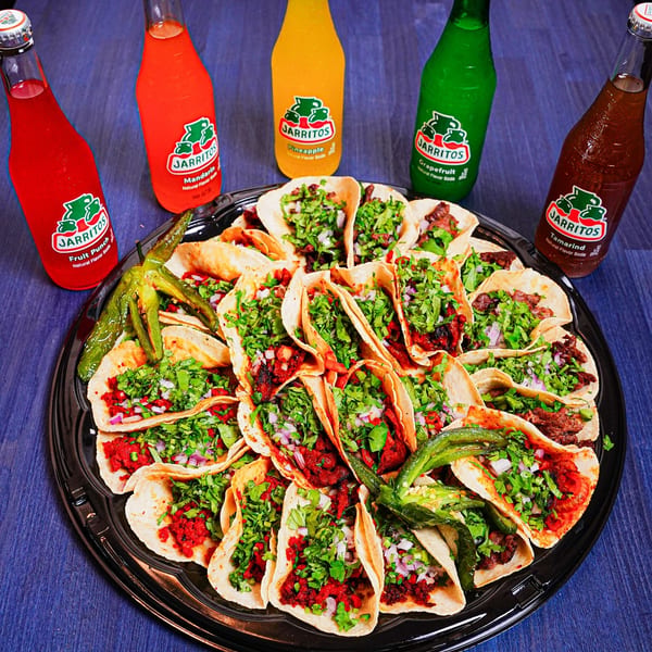 Large platter of cilantro-topped tacos with grilled peppers, surrounded by colorful Jarritos fruit soda bottles.