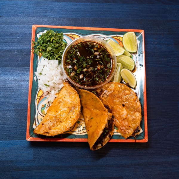 Three birria tacos with a side of consome, chopped onions, cilantro, and lime wedges on a colorful plate.