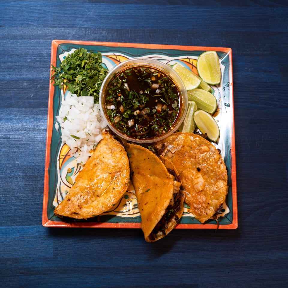 Three birria tacos with a side of consome, chopped onions, cilantro, and lime wedges on a colorful plate.