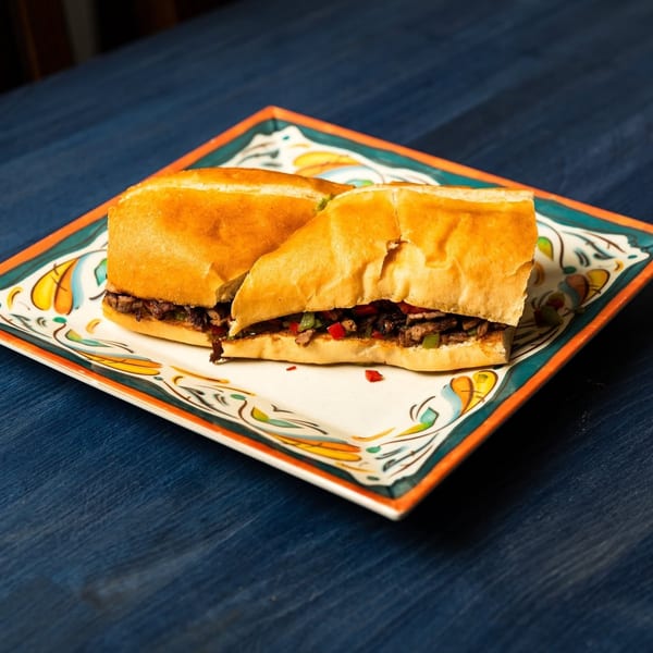 Two toasted steak sandwiches with peppers and onions on a colorful decorative plate atop a dark blue table.