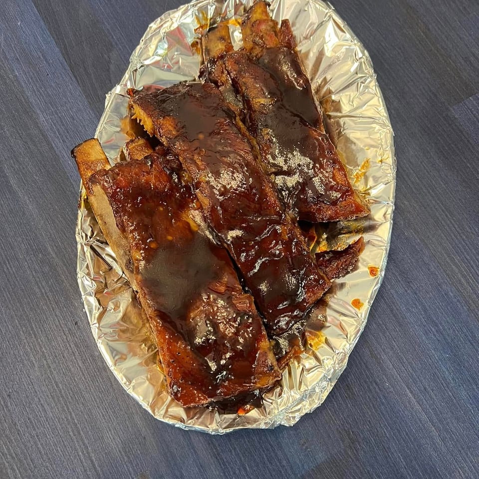Glazed barbecue ribs and grilled meat arranged on a foil-lined tray over a dark wooden table.